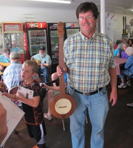 Tony Jones owns the separate farms where Tom Dula and Laura Foster are buried. Here, he holds a banjo his father had, which was made by Stanley Hicks (1980 Brown-Hudson Folklore Award recipient). At left, Austin, the fourth-generation of Grocery Basket workers, carries a canvas for guests to sign. Monday, September 7, 2015. (Leanne E. Smith)